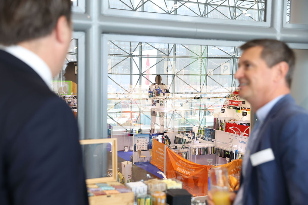 Two professionals networking at a trade show, overlooking the Texworld USA exhibition hall through large glass windows at the Javits Center